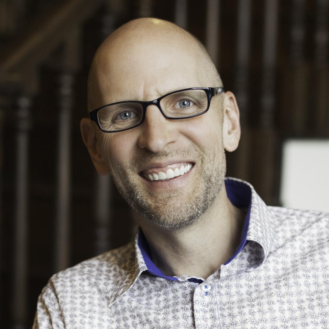 Portrait of John Farkas, smiling, in glasses and a patterned shirt