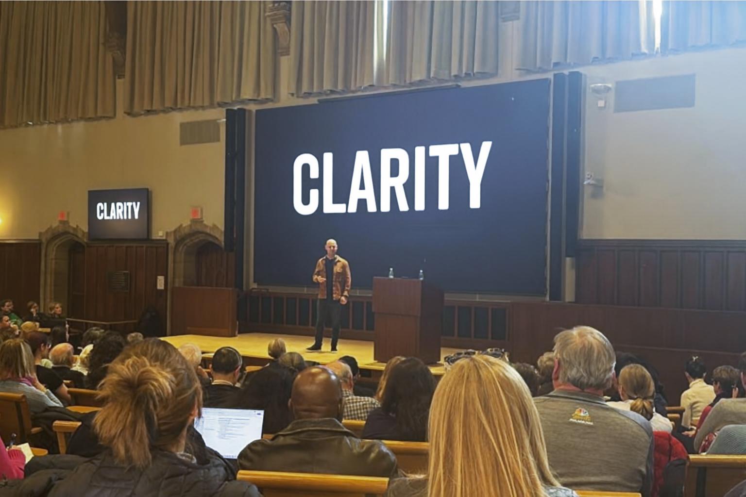 John Farkas on stage before a full auditorium, speaking in front of a large screen that reads CLARITY